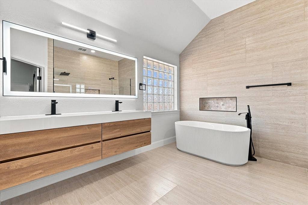 4421 Red Barn, Richardson, TX 75082 Floating double vanity with a dual-basin integrated countertop, ribbed freestanding soaking tub, matte black plumbing fixtures, large LED-backlit mirror, and full-height textured tile wall
