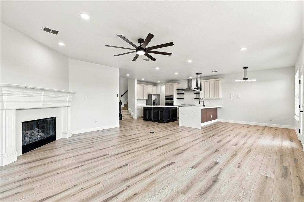 4421 Red Barn, Richardson, TX 75082 Open-concept interior featuring light wood-finish flooring, a white-mantel fireplace, a central kitchen island with a dark finish, integrated stainless steel appliances, and a multi-blade ceiling fan