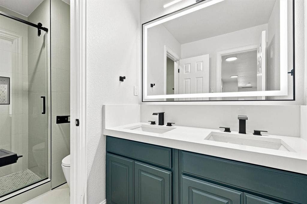 4421 Red Barn, Richardson, TX 75082 Dual-sink vanity with deep teal cabinetry and a solid-surface countertop, rectangular undermount sinks, matte black widespread faucets, and a large, integrated LED-backlit mirror