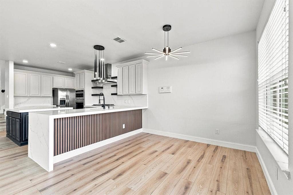 4421 Red Barn, Richardson, TX 75082 Kitchen featuring wood-finish flooring, a waterfall-edge stone countertop, vertical wood-slat island detailing, white cabinetry, and modern geometric pendant lighting