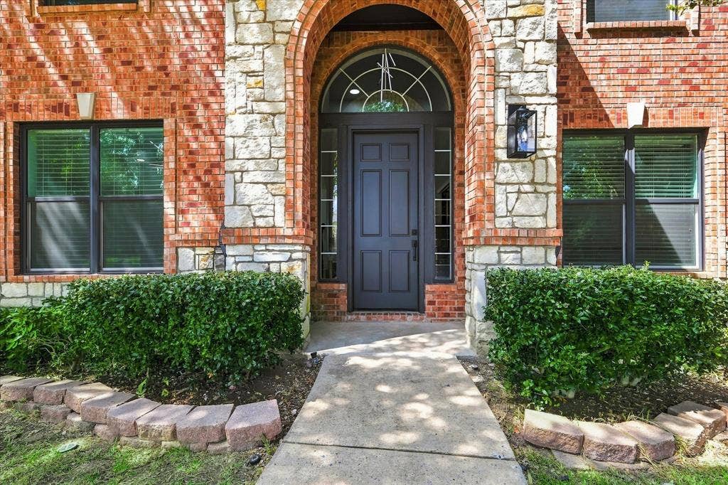 4421 Red Barn, Richardson, TX 75082 Exterior facade featuring a red brick and stone masonry veneer, arched entryway with a transom window, six-panel dark-finish entry door, and stone-bordered landscaping beds
