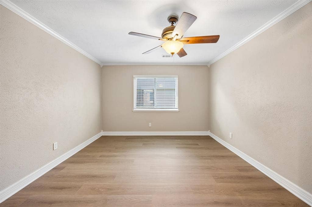 1213 Lombardy Drive, Plano, TX 75023 Enjoy the simple elegance of this inviting bedroom, featuring warm wood-look flooring, soft neutral walls, and decorative white crown molding. A window with white blinds brings in natural light, while the ceiling fan adds comfort and functionality. The elongated layout and generous wall space provide excellent versatility for a variety of uses.
