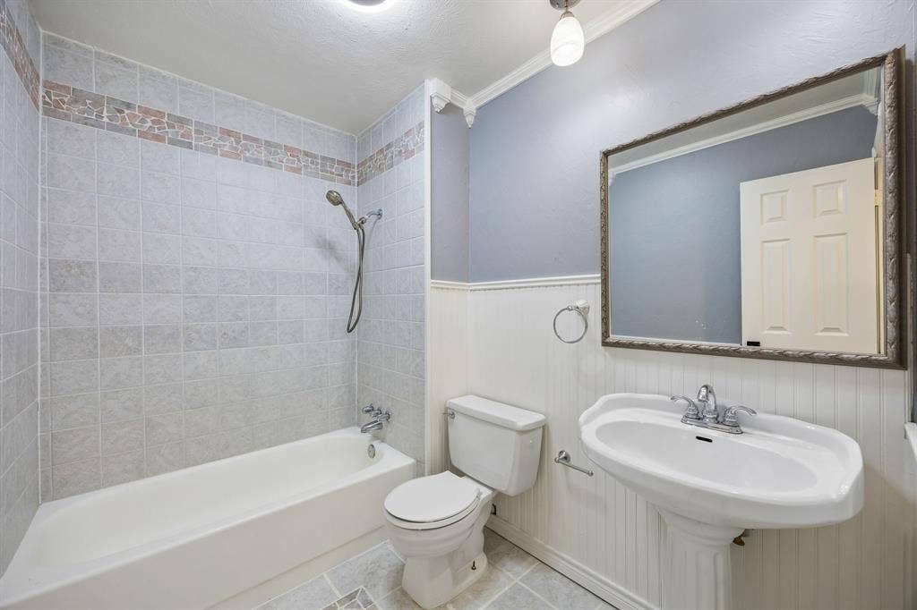 1213 Lombardy Drive, Plano, TX 75023 This well-appointed full bathroom features a classic white bathtub and shower combination surrounded by light-colored tile, complemented by a decorative accent border for added visual appeal. White beadboard wainscoting, a pedestal-style sink, and a generously sized mirror create a timeless aesthetic, while the soft blue-gray walls and coordinating tile flooring contribute to a fresh, inviting atmosphere.