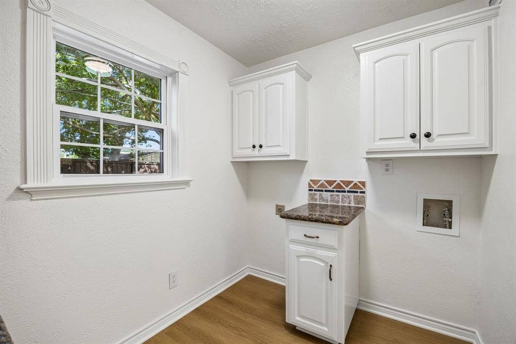 1213 Lombardy Drive, Plano, TX 75023 A charming and thoughtfully arranged utility room features white cabinetry, granite countertops, and coordinating tile accents that carry the home's distinctive design throughout. A large window welcomes natural light and offers a pleasant view of the outdoors, while the dedicated washer and dryer connections provide a convenient space for everyday laundry needs. Warm wood-look flooring and classic trim complete this bright, functional area.