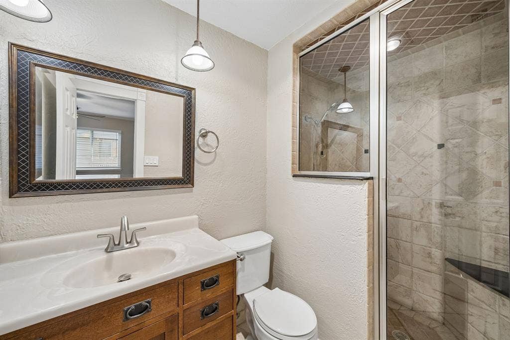 1213 Lombardy Drive, Plano, TX 75023 A well-appointed bathroom features a warm wood-tone vanity with ample drawer storage, a light countertop, and coordinating fixtures. The expansive framed mirror enhances the sense of space, while the tiled walk-in shower showcases a decorative tile pattern, glass enclosure, and built-in bench. Neutral finishes and thoughtful details create a comfortable, functional space.