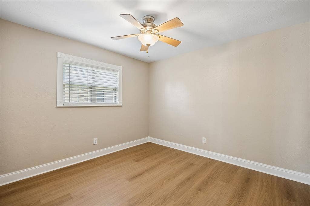 1213 Lombardy Drive, Plano, TX 75023 A bright and inviting bedroom showcases warm wood-look flooring, soft neutral walls, crisp white trim, and a charming ceiling fan with integrated lighting. A well-positioned window with white blinds welcomes natural light, creating a comfortable and versatile space ready to suit a variety of needs.