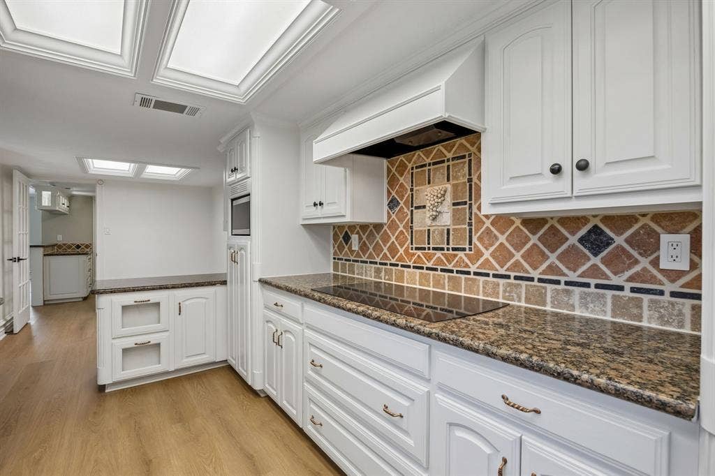 1213 Lombardy Drive, Plano, TX 75023 Another view of the beautifully appointed kitchen showcasing crisp white cabinetry with decorative molding, rich granite countertops, and a distinctive multicolored tile backsplash that adds warmth and character. A sleek cooktop, built-in microwave, and custom range hood create a functional cooking space, while recessed ceiling panels and abundant cabinetry enhance the room's timeless appeal. Warm wood-look flooring flows throughout, complementing the inviting neutral palette.