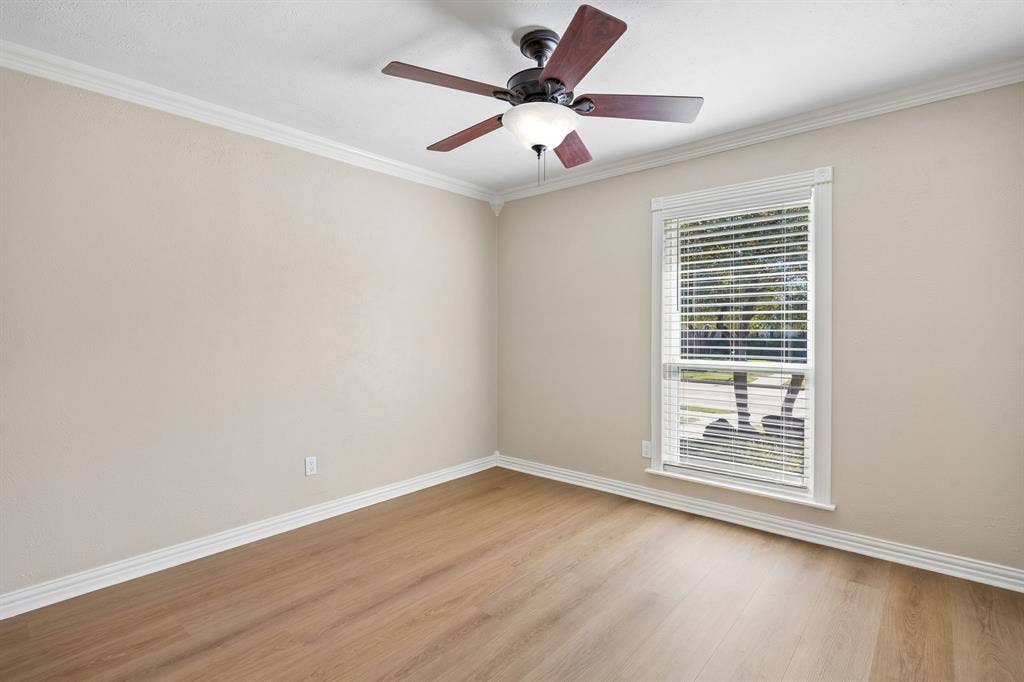 1213 Lombardy Drive, Plano, TX 75023 A comfortable bedroom offers a peaceful retreat with soft neutral walls, warm wood-look flooring, crisp white baseboards, and elegant crown molding. A ceiling fan with integrated lighting adds everyday comfort, while the generously sized window welcomes natural light and provides views of the surrounding greenery. The clean lines and versatile color palette create an inviting canvas for a variety of design styles.