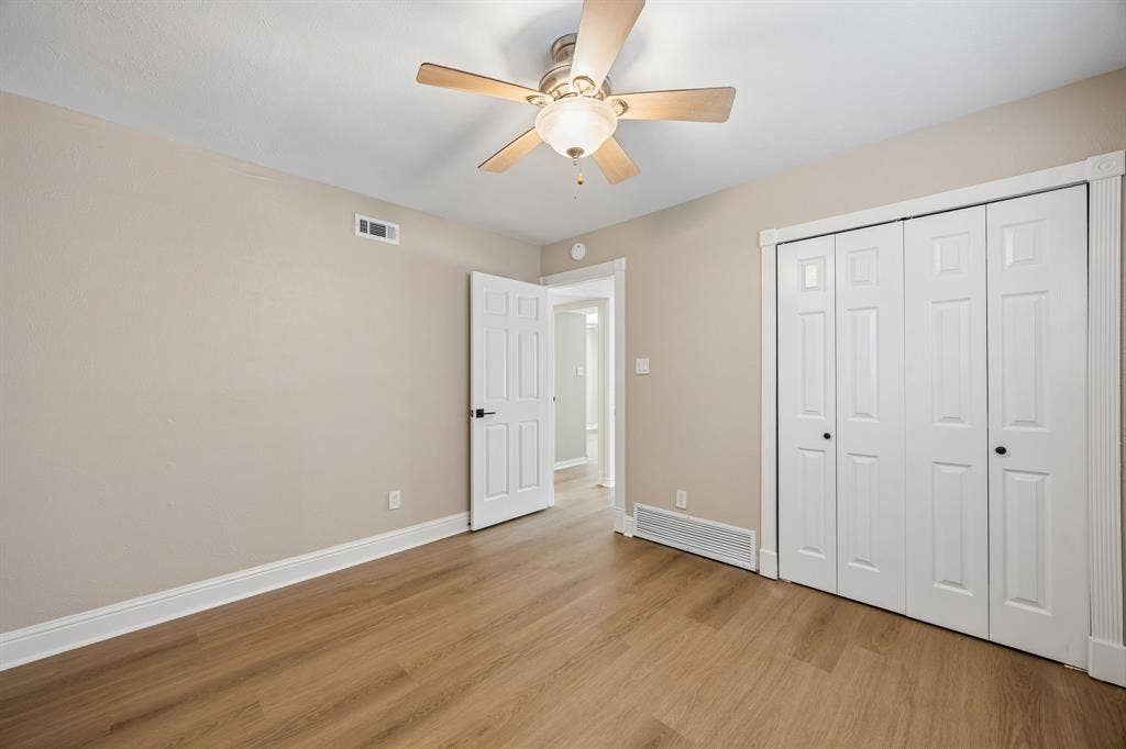 1213 Lombardy Drive, Plano, TX 75023 This spacious bedroom features beautiful wood-look flooring, soothing neutral walls, and elegant white trim that creates a clean, cohesive look. A generous double-door closet provides convenient storage, while the ceiling fan and overhead lighting add everyday comfort. The open doorway offers an inviting transition to the adjoining living spaces.