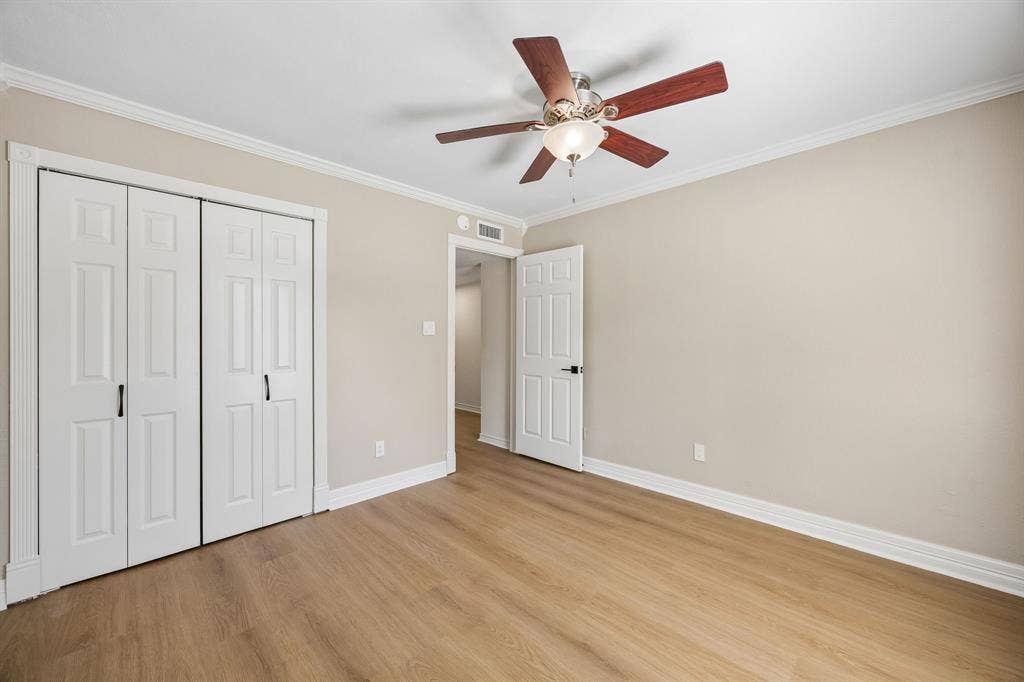 1213 Lombardy Drive, Plano, TX 75023 A spacious bedroom showcases warm wood-look flooring, soft neutral walls, and classic white architectural trim that lends a polished, cohesive finish. A ceiling fan with integrated lighting adds comfort, while the wide closet with traditional paneled doors offers practical storage. The generous open floor area and understated color scheme create a flexible space ready to accommodate a variety of uses and design preferences.