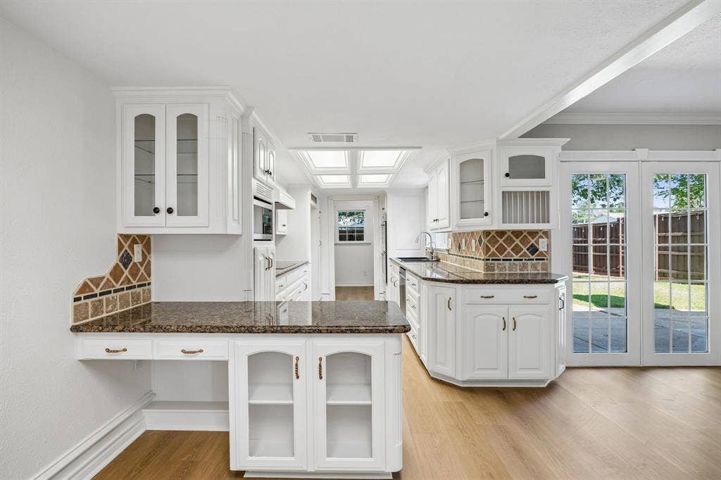 1213 Lombardy Drive, Plano, TX 75023 A bright, thoughtfully arranged kitchen features abundant white cabinetry, richly patterned countertops, and distinctive diamond-pattern tile backsplash detailing. A central peninsula provides additional workspace and storage, while glass-front cabinet doors and decorative millwork add character. Overhead natural-light panels brighten the space, and French doors offer convenient access to the backyard.