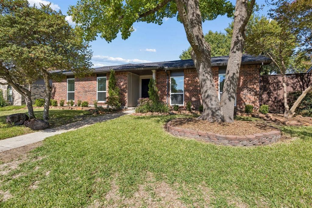 1213 Lombardy Drive, Plano, TX 75023 Mature trees provide a picturesque canopy over the front lawn, complementing the home's warm brick facade and established landscaping. A graceful walkway leads to the covered entrance, while a raised stone garden bed adds character and dimension to the beautifully shaded front yard.