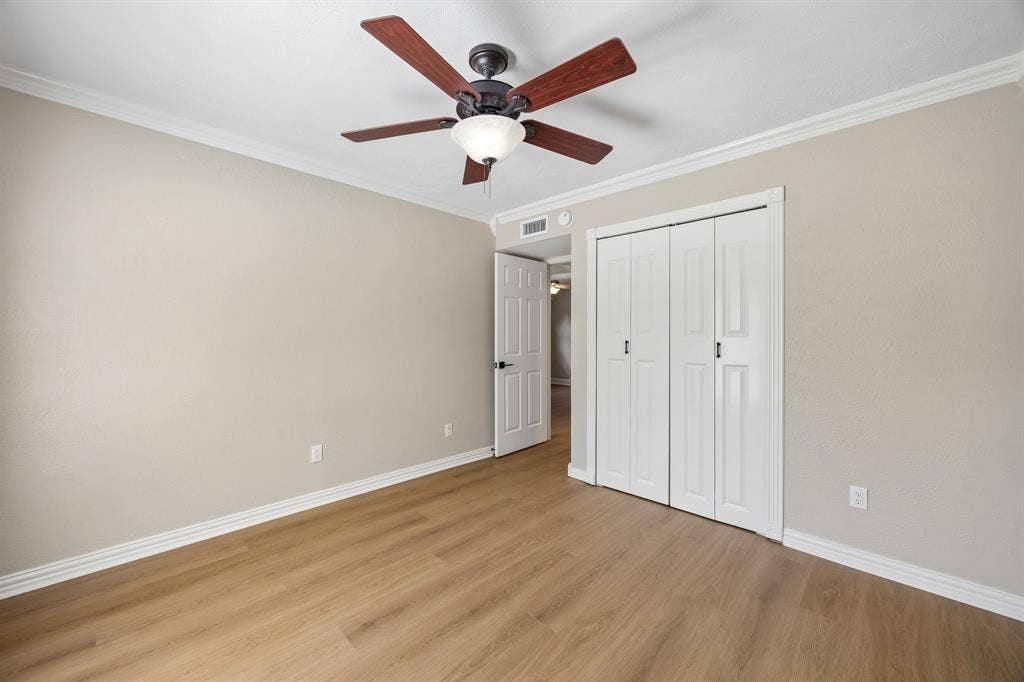 1213 Lombardy Drive, Plano, TX 75023 This inviting bedroom combines understated elegance with practical features, including warm wood-look flooring, decorative crown molding, and a ceiling fan with integrated lighting. A spacious closet with classic white folding doors provides convenient storage, while the neutral finishes and open floor area allow flexibility for furniture placement. The bright, uncluttered presentation highlights the room's comfortable proportions and timeless appeal.