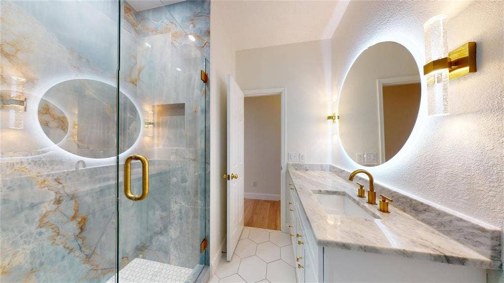 10514 Greenbriar Lane, Rowlett, TX 75089 Bathroom featuring a walk-in shower with large-format decorative wall tile, brass-finish hardware, a stone countertop vanity with integrated sink, a backlit circular mirror, and hexagonal floor tile