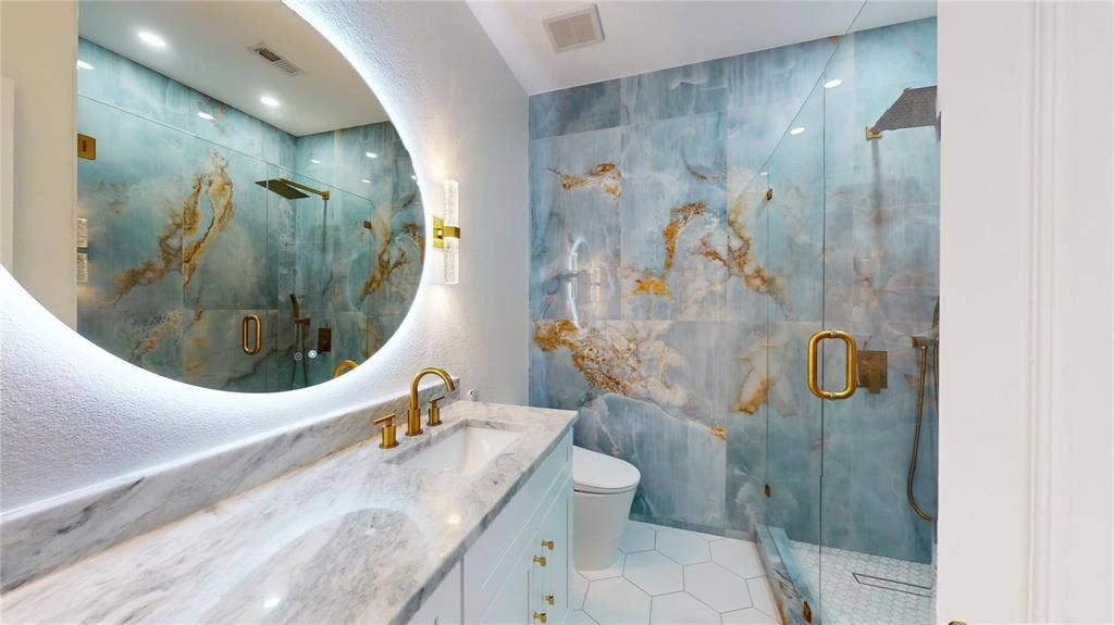 10514 Greenbriar Lane, Rowlett, TX 75089 Bathroom featuring large-format blue and gold-toned wall tile, a frameless glass-enclosed shower, a backlit circular mirror, brass-finish hardware, and a stone-surface vanity