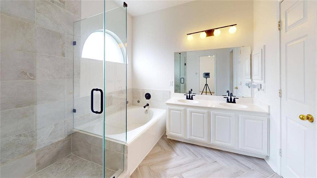 10514 Greenbriar Lane, Rowlett, TX 75089 Dual-sink vanity with white cabinetry, glass-enclosed shower with tile surround, soaking tub, arched window, and herringbone-patterned tile flooring