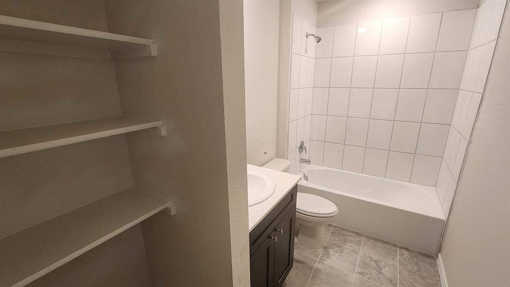 Built-in shelving, dark wood-finish vanity with countertop, undermount sink, white tile shower surround, and gray-toned floor tiling