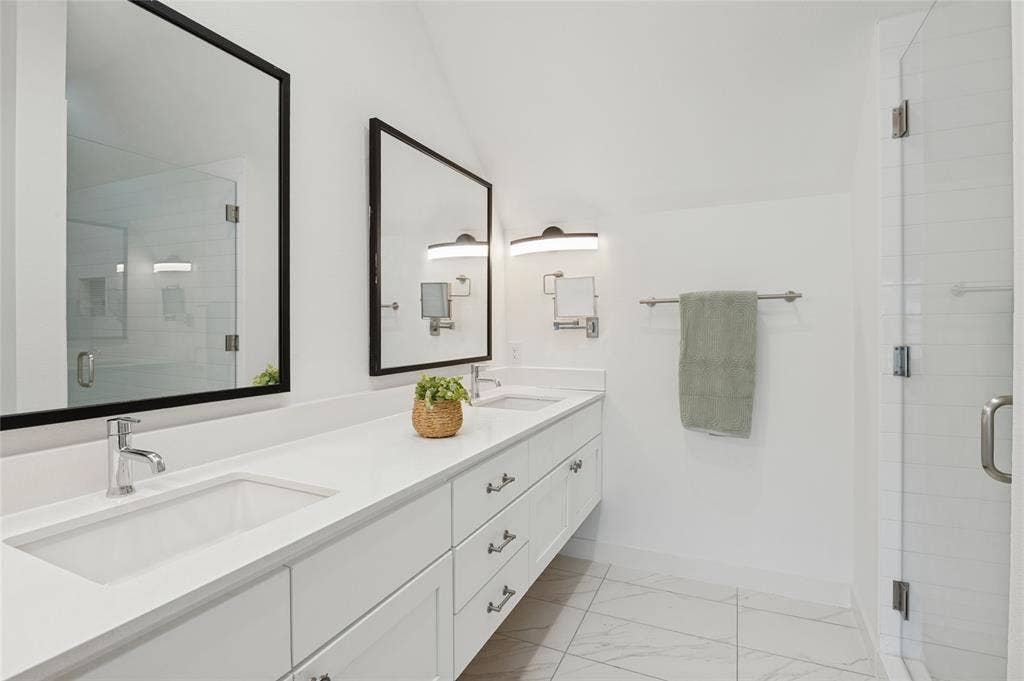 614 Finley Court, Dallas, TX 75208 Dual-sink vanity with a solid-surface countertop, white cabinetry with metal hardware, framed mirrors, wall-mounted lighting, and a glass-enclosed shower with tiled flooring