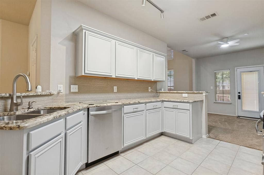 Kitchen featuring white shaker-style cabinetry, neutral stone-patterned countertops, a stainless steel undermount double-basin sink with a gooseneck faucet, light-toned tiled flooring, and integrated stainless steel dishwasher