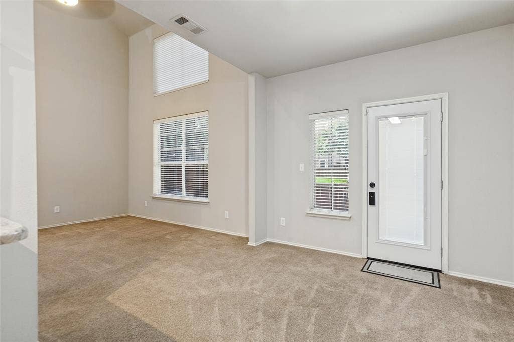 High-ceilinged room featuring neutral wall tones, carpeted flooring, multiple oversized windows, and a glass-paneled exterior entry door