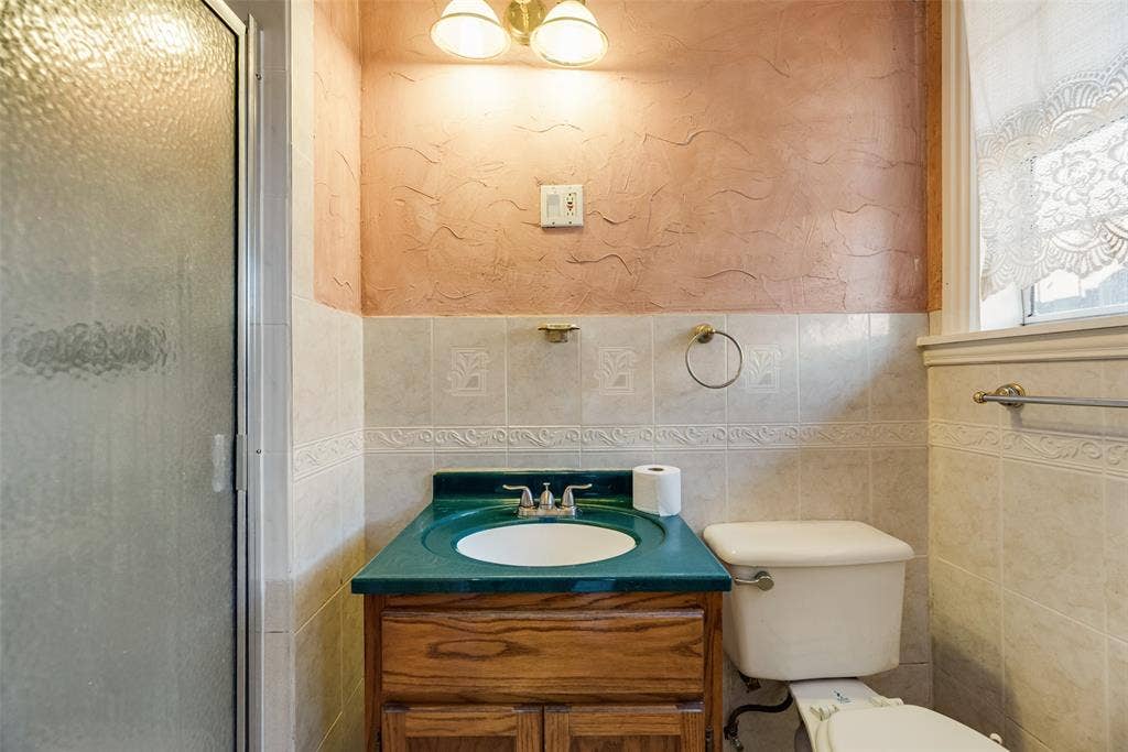 1572 Gillette Circle, Dallas, TX 75217 Bathroom featuring a wood-finish vanity with an integrated basin, textured wall finishes, a tiled backsplash with decorative inlay, and a glass-enclosed shower