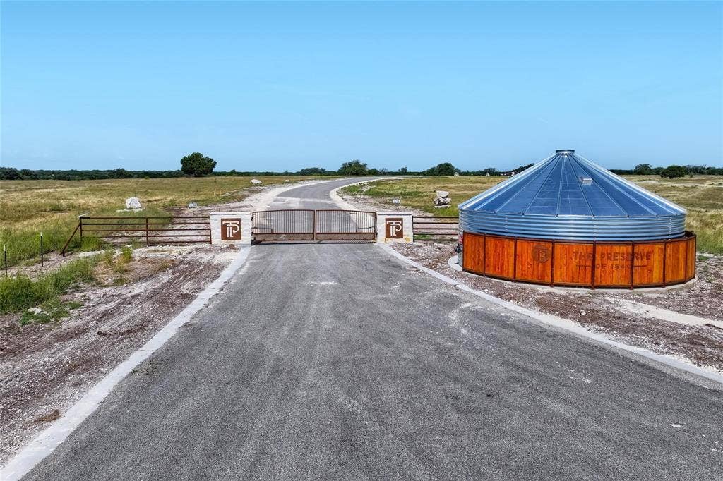 Gated entrance featuring masonry pillars, metal security gate, paved driveway, and custom circular water storage structure with wood-finish siding and metal roofing
