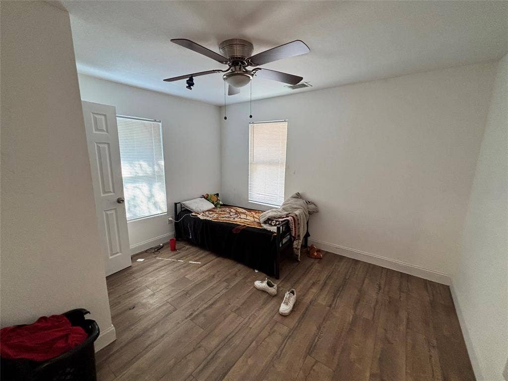 Neutral interior room featuring wood-finish flooring, two windows with blinds, a multi-panel interior door, and a ceiling fan with integrated light fixture