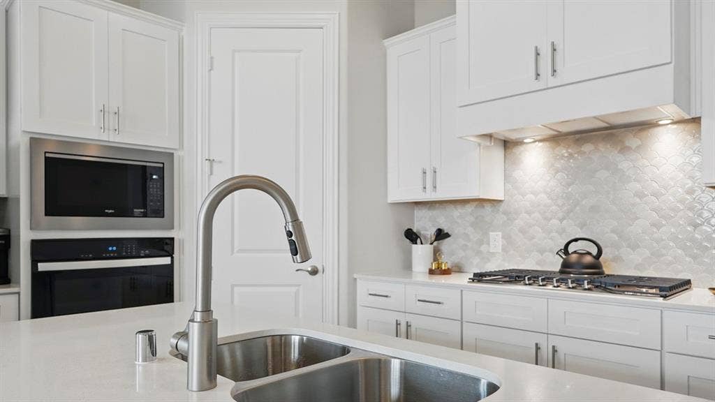 White cabinetry, stone-finish countertops, stainless steel dual-basin sink, scalloped tile backsplash, and built-in stainless steel double wall oven and microwave