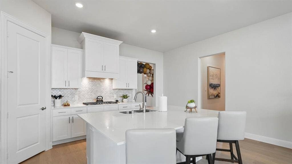 Kitchen featuring white shaker-style cabinetry, a stone-surfaced island with an integrated sink, geometric tile backsplash, and wood-finish flooring