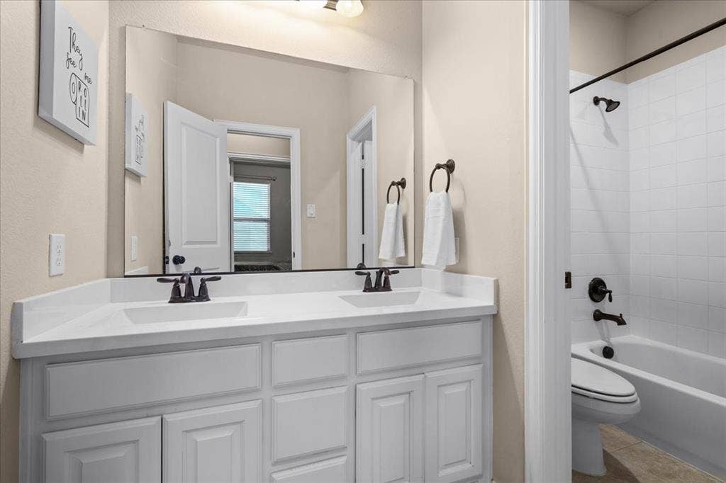 Dual-sink vanity with white cabinetry, solid-surface countertop, bronze fixtures, large vanity mirror, and a tub-shower combination featuring white tile surround