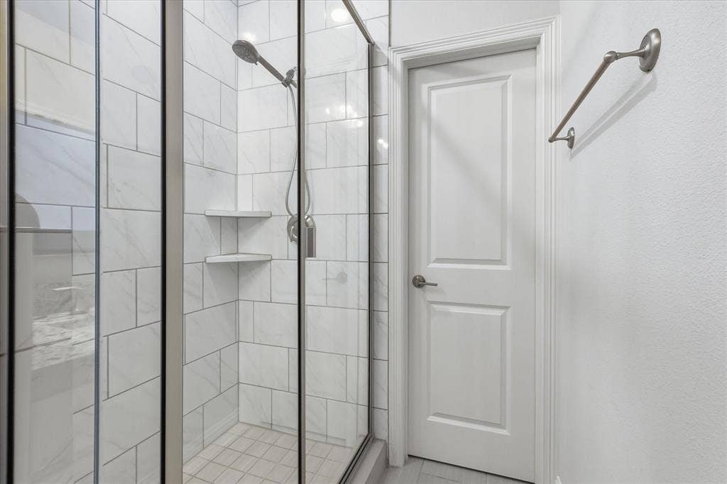 Walk-in shower featuring a glass enclosure, wall-mounted showerhead, built-in corner shelving, and tile-surround walls, complemented by a solid-core paneled door and metal wall-mounted towel bar
