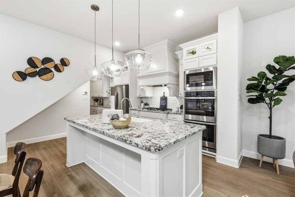 Kitchen featuring a central island with stone countertop, stainless steel double wall ovens, white cabinetry, decorative pendant lighting, and wood-finish flooring