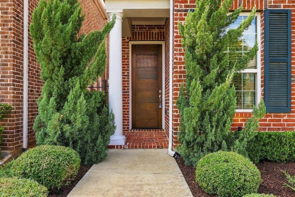 Brick exterior facade featuring a horizontal wood-paneled front door, classic columned entryway, decorative shutters, and landscaped greenery