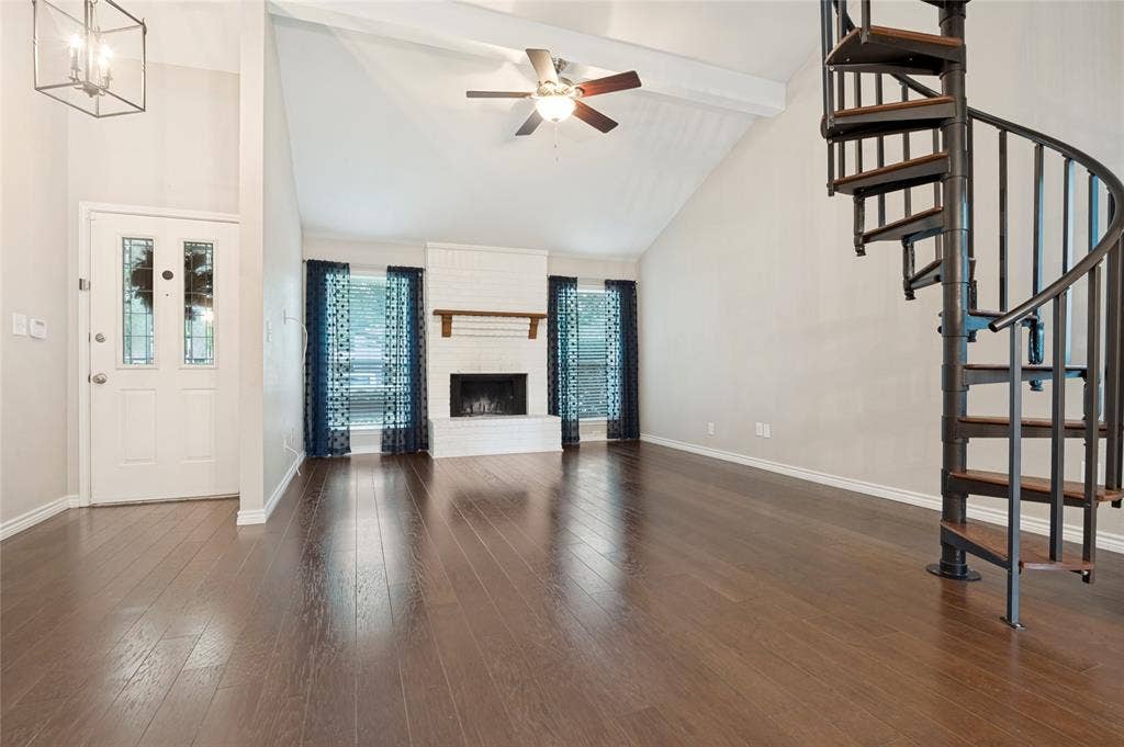 Vaulted ceiling with exposed beam and ceiling fan, dark wood-finish flooring, white brick fireplace with wood mantel, leaded glass front door with transoms, black metal spiral staircase