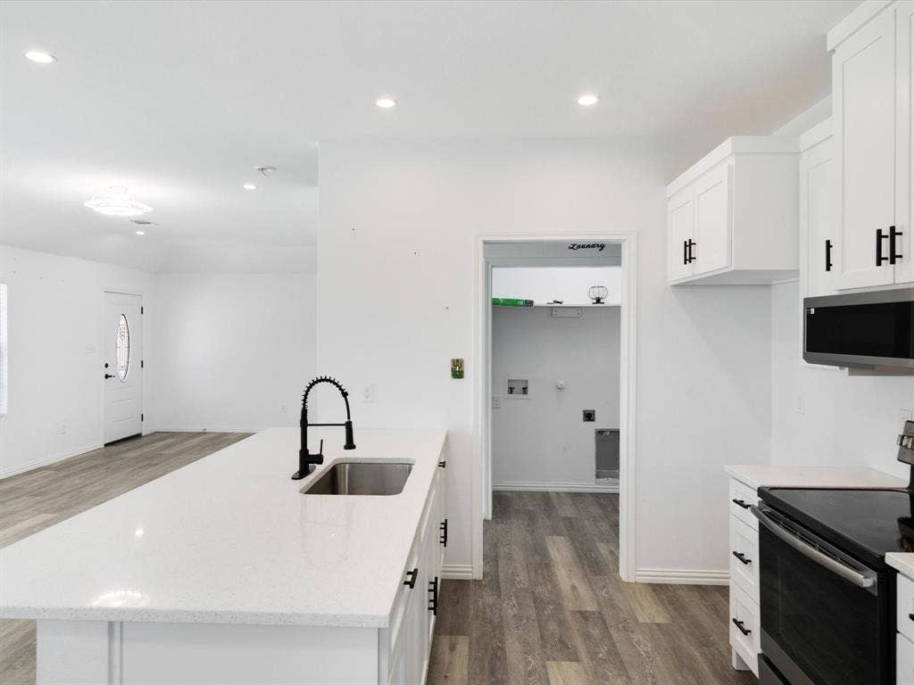 Kitchen featuring a central island with a stone-finish countertop and undermount stainless steel sink, white shaker-style cabinetry, matte black hardware, recessed lighting, and wood-finish flooring