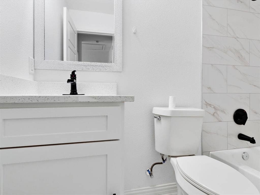 Bathroom featuring a white vanity cabinet, speckled stone-finish countertop, matte black faucet and shower fixtures, rectangular wall-mounted mirror, and tile surround with a marble-patterned finish