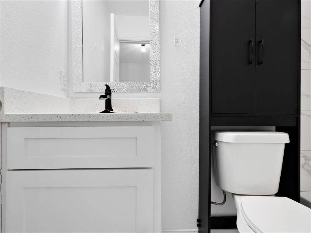 White shaker-style vanity with a light-toned speckled countertop, matte black faucet, framed mirror, and a black over-the-toilet storage unit