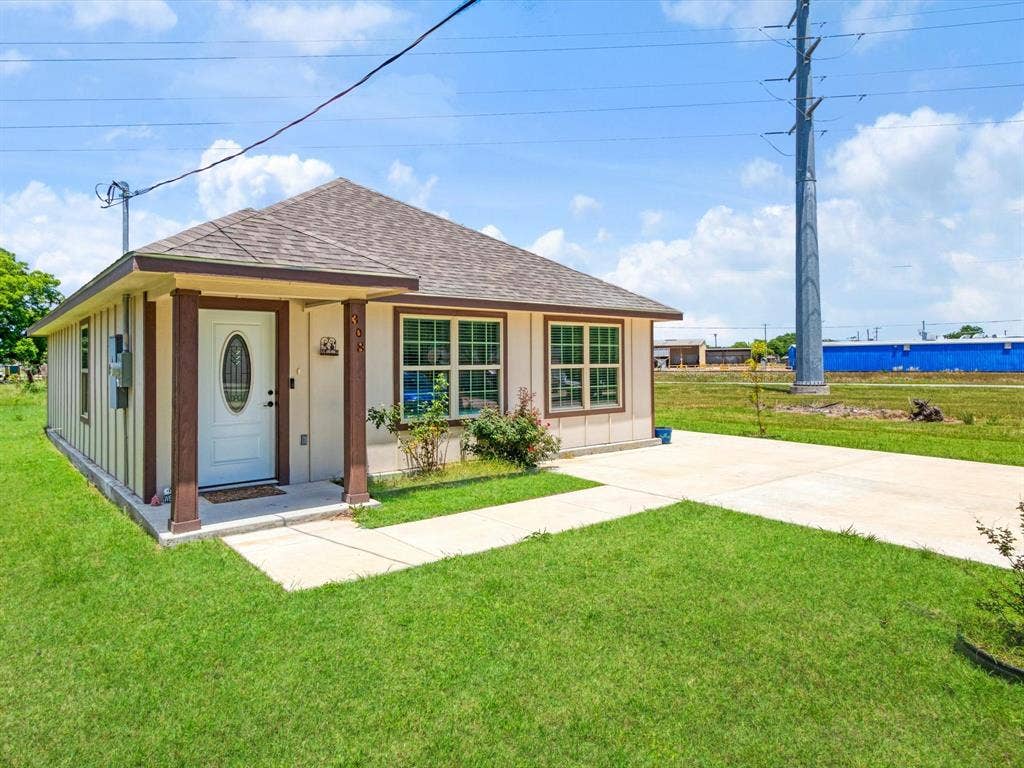 Single-story exterior featuring a shingled hipped roof, paneled siding, covered front porch with support pillars, and a concrete driveway