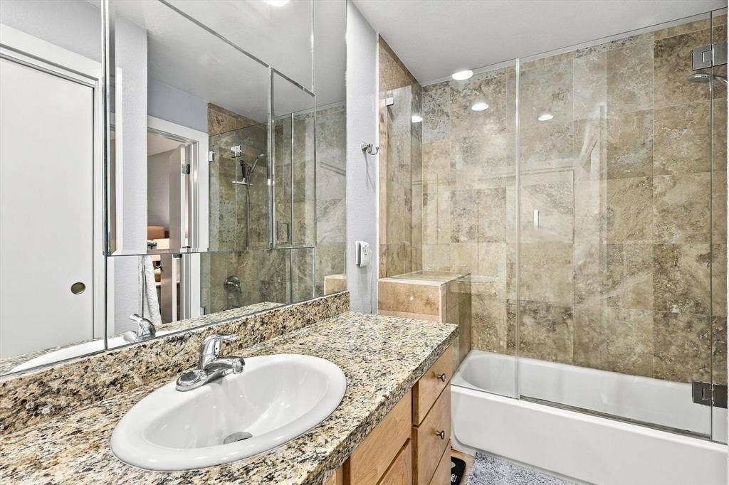 4777 Cedar Springs Road, #9K, Dallas, TX 75219 Bathroom featuring a vanity with stone countertop, an integrated oval sink, full-width mirror, and a tub-shower combination enclosed by a glass panel with floor-to-ceiling stone-tile surround