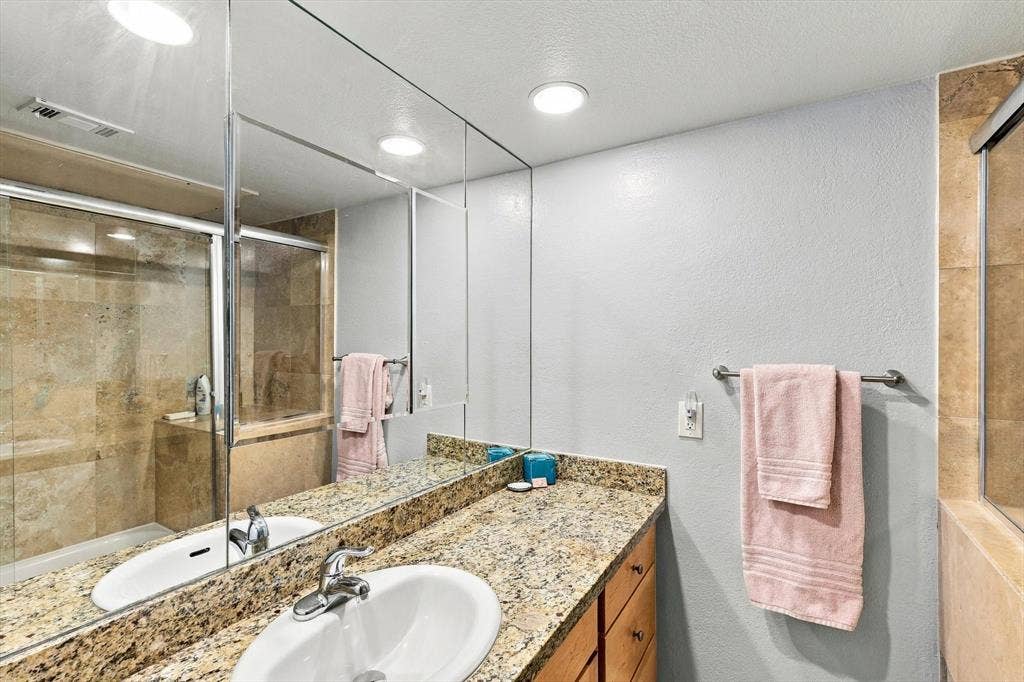 4777 Cedar Springs Road, #9K, Dallas, TX 75219 Full-width wall mirror, stone-surfaced vanity top with an integrated oval sink, wood-finish cabinetry, and a walk-in shower enclosure featuring tiled walls and sliding glass doors