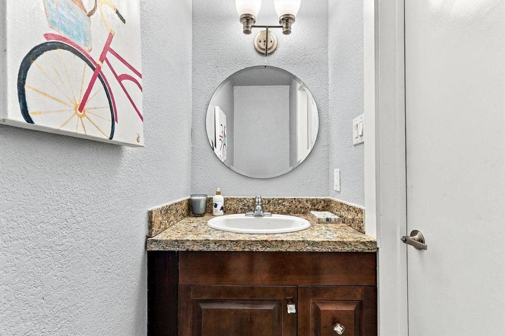 4777 Cedar Springs Road, #9K, Dallas, TX 75219 Bathroom featuring a dark wood cabinet, stone-surface countertop with an integrated basin, circular wall-mounted mirror, and dual-bulb overhead lighting