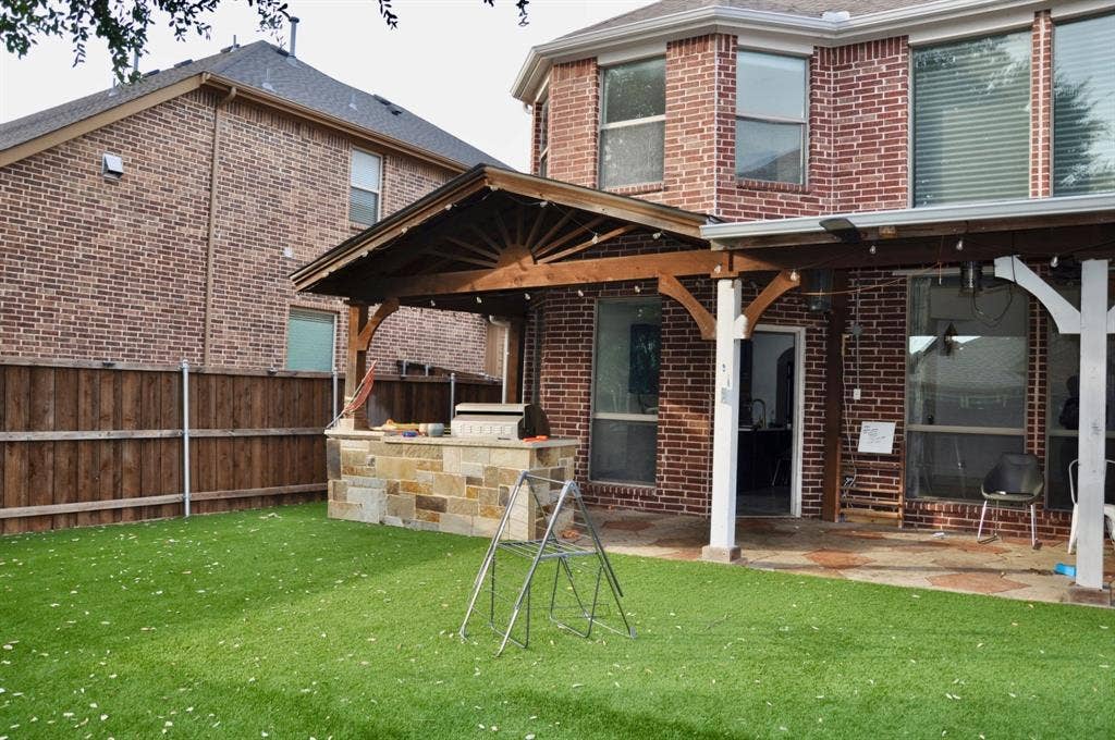 3620 Carlton Court, Sachse, TX 75048 Backyard featuring a covered patio with timber framing, stone-clad outdoor kitchen, brick exterior, and synthetic turf lawn