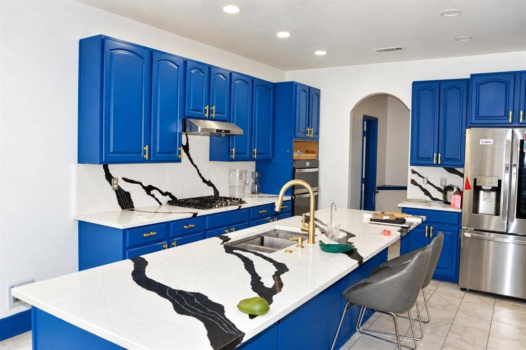 3620 Carlton Court, Sachse, TX 75048 Kitchen featuring bold blue cabinetry, veined stone-finish countertops with matching backsplash, stainless steel appliances, a center island with an integrated dual-basin sink, and brass-finish hardware