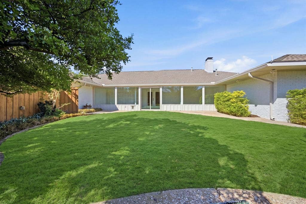 6978 Pemberton Drive, Dallas, TX 75230 Expansive backyard featuring a manicured lawn, white brick exterior, floor-to-ceiling windows, a stone patio, and a wood privacy fence