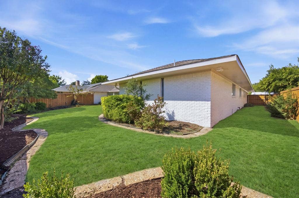 6978 Pemberton Drive, Dallas, TX 75230 White brick exterior featuring a hip roof, expansive lawn, stone-border garden beds, and wood privacy fencing