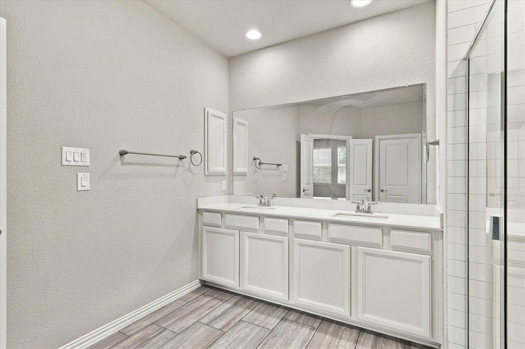 614 Ansley Way, Allen, TX 75013 Dual-vanity cabinetry with white finish, solid surface countertop, expansive wall-mounted mirror, recessed lighting, and wood-finish flooring