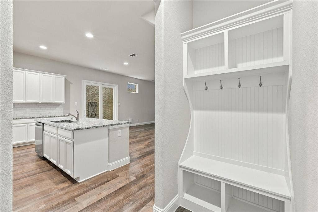 614 Ansley Way, Allen, TX 75013 Built-in white mudroom bench featuring beadboard detailing, upper storage cubbies, and garment hooks, complemented by an open-concept kitchen area with white cabinetry, stone-finish countertops, and wood-finish flooring