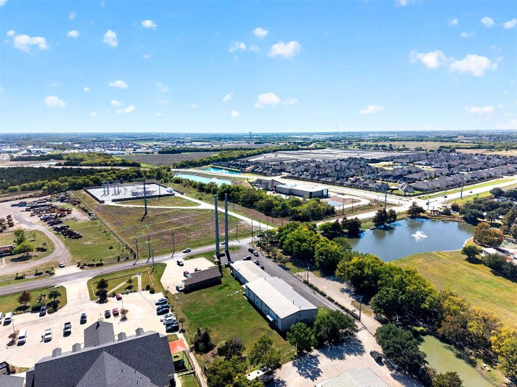 911 W Holiday Drive, Fate, TX 75132 Aerial perspective showcasing expansive acreage, a scenic pond with a fountain, mature tree clusters, and direct access to paved thoroughfares