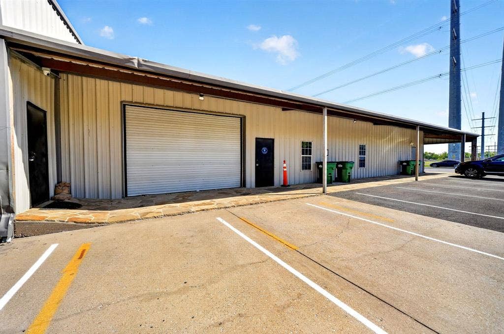 911 W Holiday Drive, Fate, TX 75132 Metal-sided commercial exterior featuring a roll-up garage door, stone walkway, multiple access doors, and dedicated asphalt parking