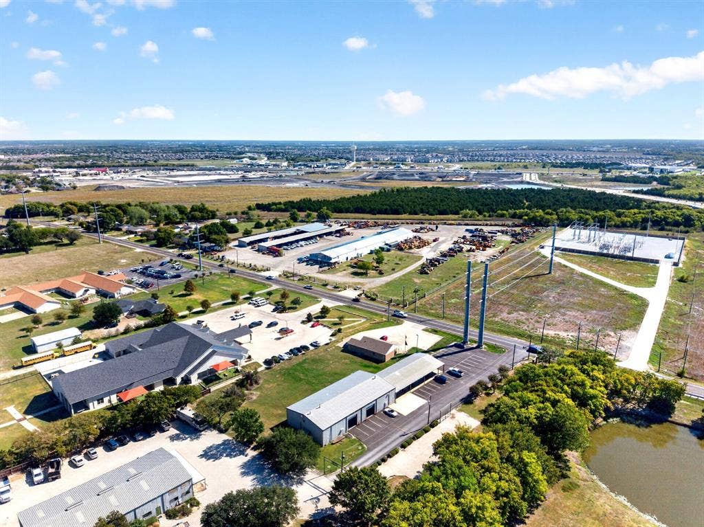 911 W Holiday Drive, Fate, TX 75132 Aerial perspective of a multi-structure commercial property featuring expansive asphalt parking areas, covered storage bays, metal-roofed industrial buildings, and direct access to paved roadways